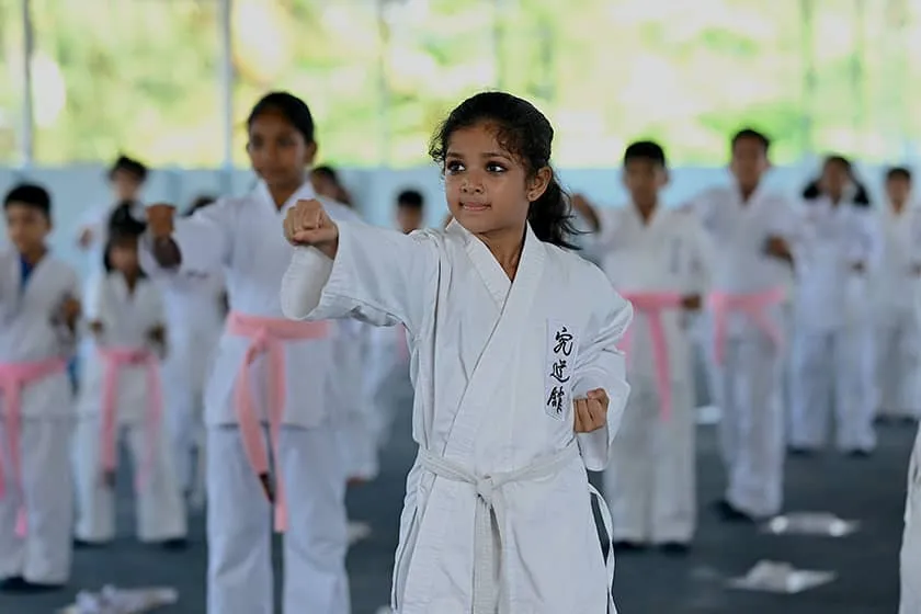 A young karateka in a white gi practising a stance with classmates behind her